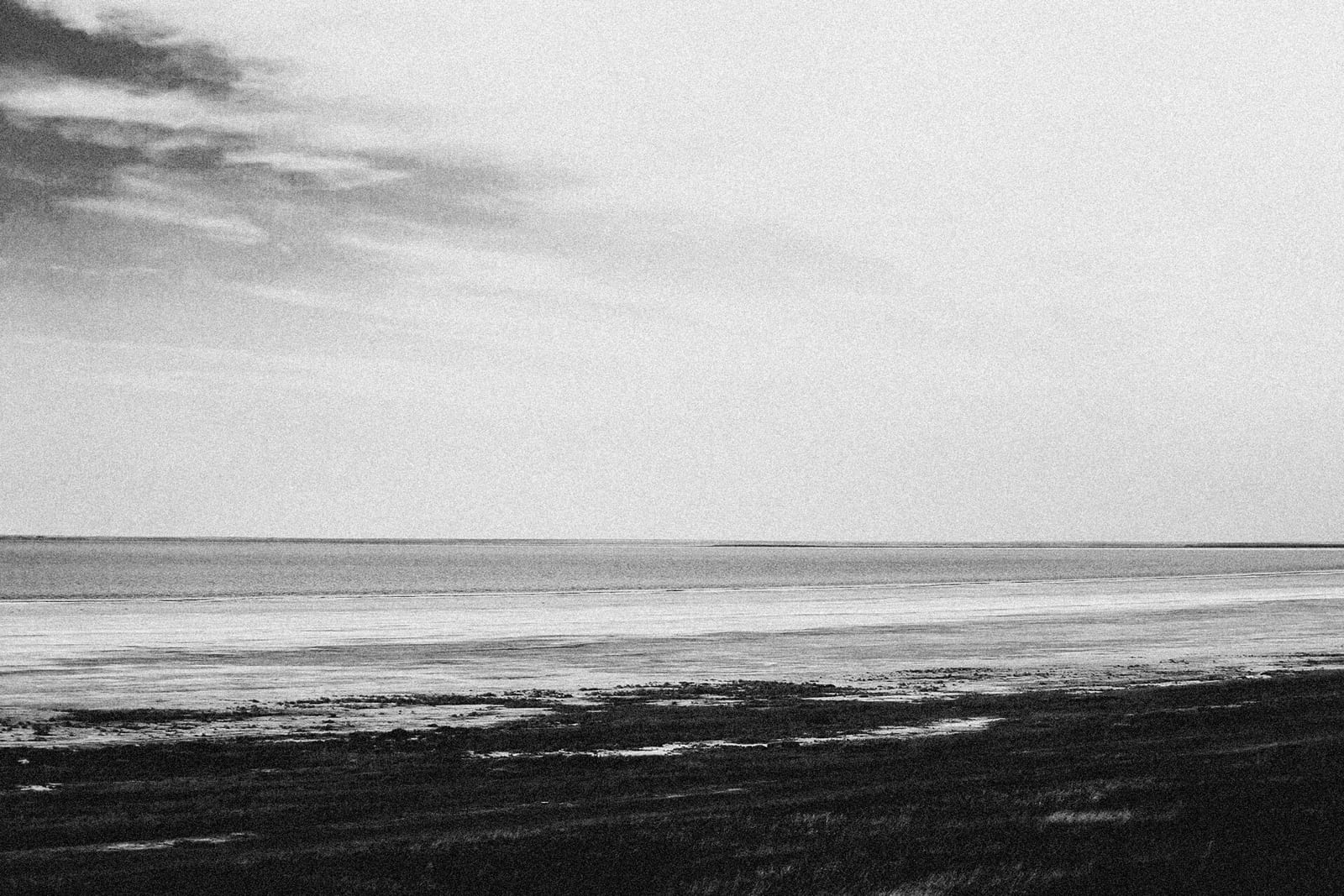 A grainy black and white photograph of a wide, empty shoreline under a pale sky.